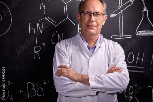 elderly caucasian scientist man posing near blackboard with formulas, looks at camera, in white medical wear and eyeglasses. science, chemistry