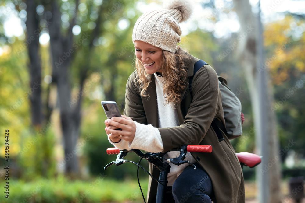 Beautiful young woman sending messages with her smart phone while cycling through the park in autumn. Beautiful young woman sending messages with her smart phone while cycling through the park in autumn.