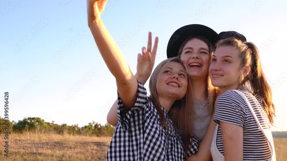 Three cheerful young women take pictures of their front camera on a field at sunset. Cheerful girls take selfies and smile in front of the camera. Three cheerful young women take pictures of their front camera on a field at sunset. Cheerful girls take selfies and smile in front of the camera.