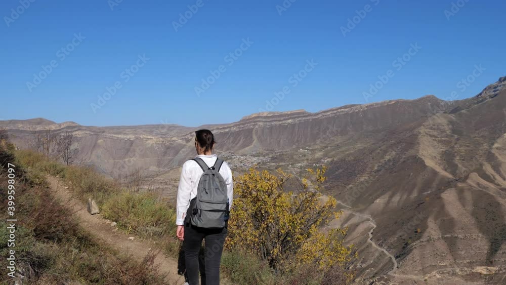 Young woman in a white shirt with a backpack climbs the trail against the background of the mountains on an autumn clear day.