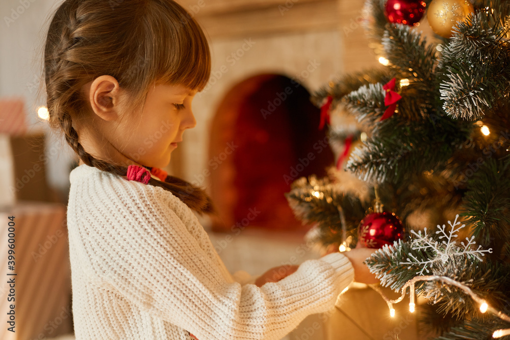 Kid decorating Christmas tree, charming girl with pigtails wearing white sweater, looking at fur tree with garland, posing in festive room, child waits New year tree. Kid decorating Christmas tree, charming girl with pigtails wearing white sweater, looking at fur tree with garland, posing in festive room, child waits New year tree.