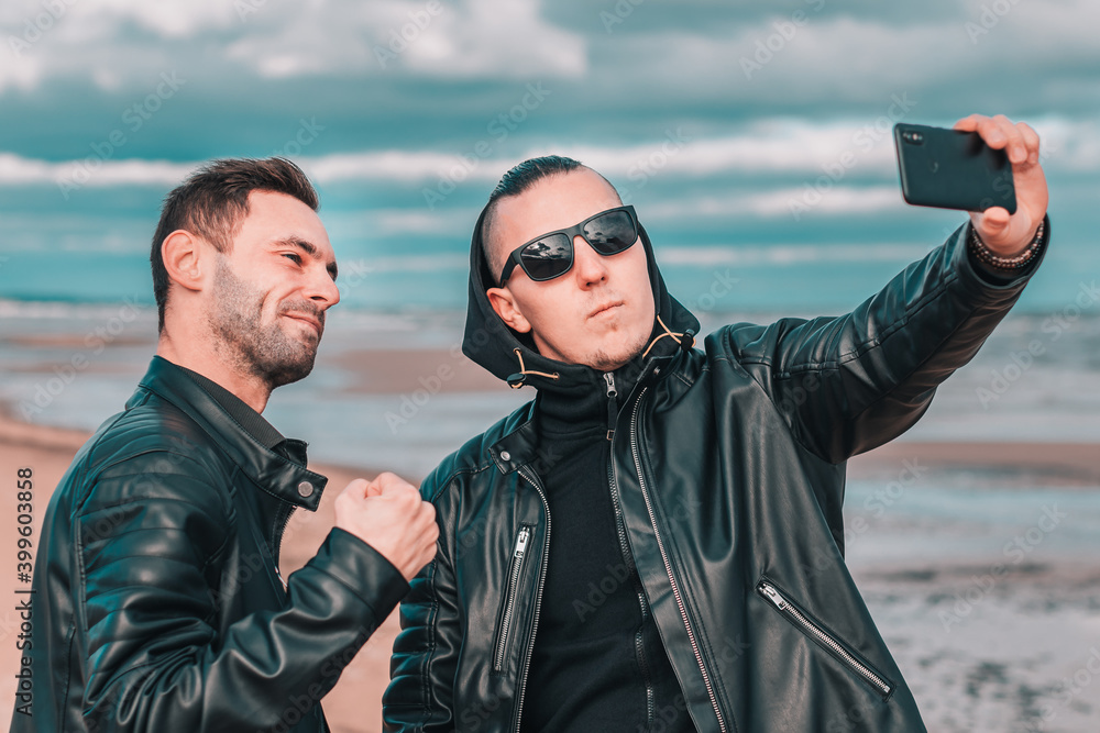 Two Handsome Male Friends Making Selfie Using Black Smartphone at the Beach. Youthful Men in Black Clothes Having Fun by Making Photos Against the Sea Two Handsome Male Friends Making Selfie Using Black Smartphone at the Beach. Youthful Men in Black Clothes Having Fun by Making Photos Against the Sea