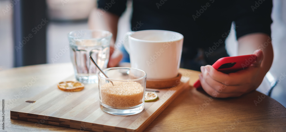Female hands with a cup of coffee and a mobile phone close-up, sugar bowl in the foreground adding sugar Female hands with a cup of coffee and a mobile phone close-up, sugar bowl in the foreground adding sugar