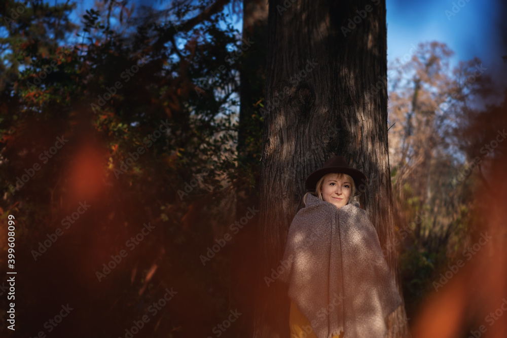 Naklejka premium A beautiful young woman in a brown felt hat stands by a tree in the forest wrapped in plaid