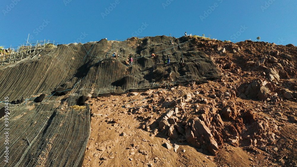 Workers strengthen the slope of the mountain with metal mesh preventing rockfall and landslide on the road, above view. workers constructing anti-landslide concrete wall prevent protect against rock