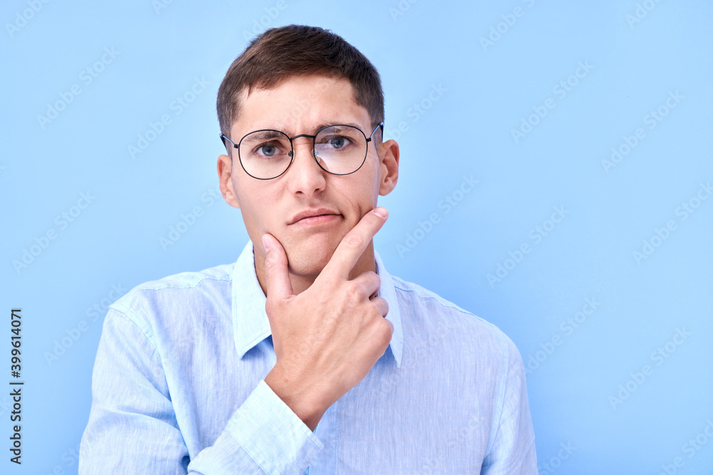 Portrait of pensive man in glasses and shirt holding his chin and thinks, makes a decision, doubts isolated in blue studio with copy space