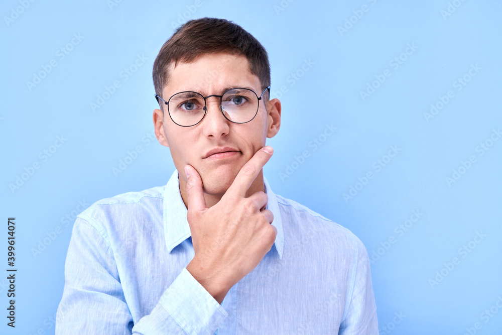 Portrait of pensive man in glasses and shirt holding his chin and thinks, makes a decision, doubts isolated in blue studio with copy space Portrait of pensive man in glasses and shirt holding his chin and thinks, makes a decision, doubts isolated in blue studio with copy space