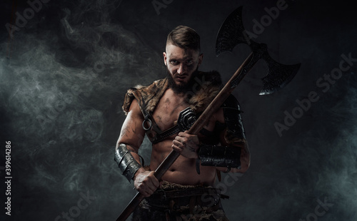 Northern vandal seafarer with muscular build holding huge axe and staring at camera in dark smokey background.