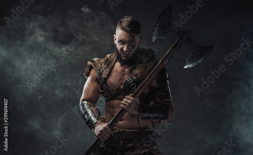 Brutal nordic heathen dressed in light armour posing in dark smokey background with huge axe and staring at camera.