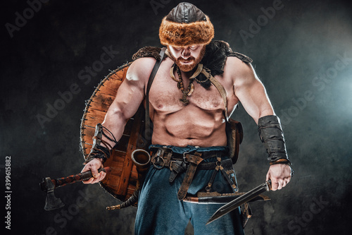 Photography Strong and sad viking warrior with beard and hat looking down in foggy background holding hatchet and knife