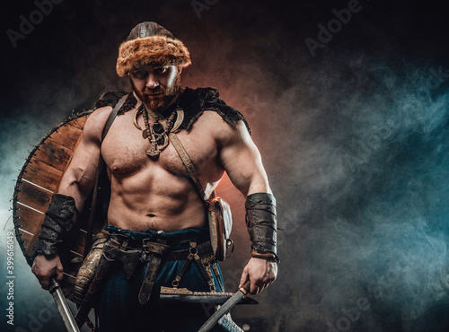 Фотография Mad and savage northern warrior with naked torso and shield on his back poses in foggy background staring at camera and holding hatchet and knife