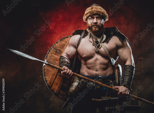 Nude handsome northern barbarian with naked torso and muscular build poses holding a spear in dark red background.