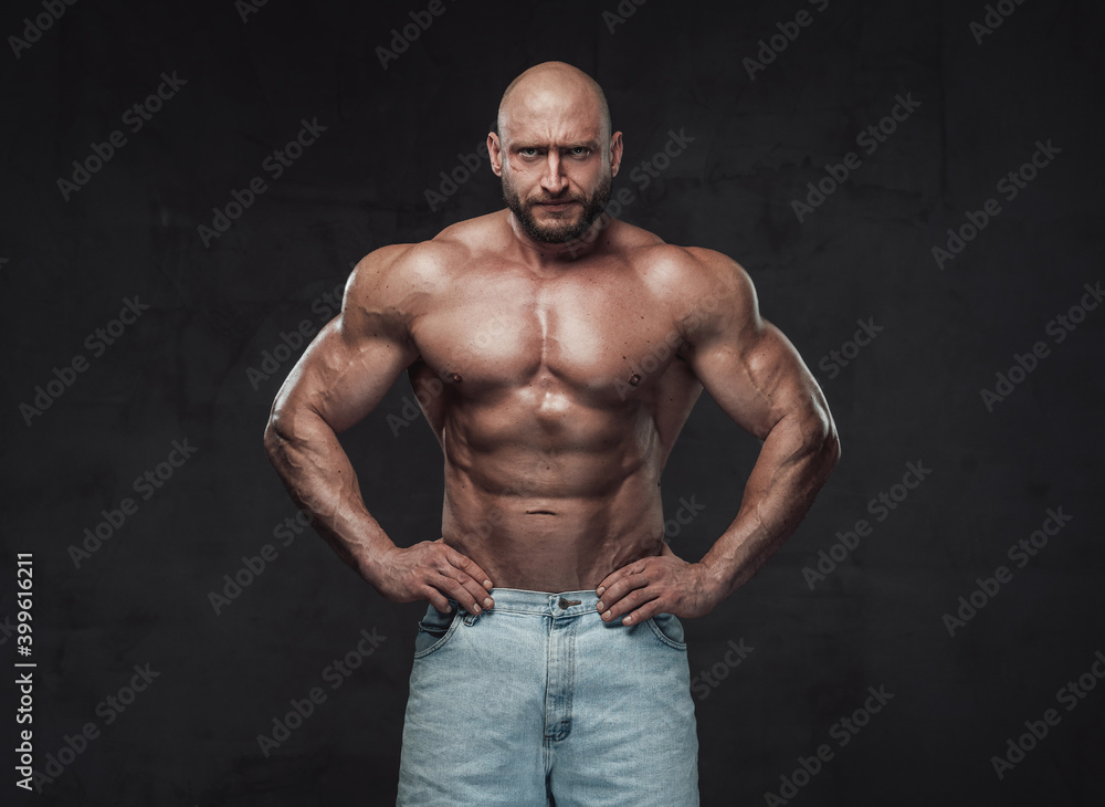 Powerful and handsome bodybuilder with hairless head poses in dark background with naked torso with angry face looking at camera.