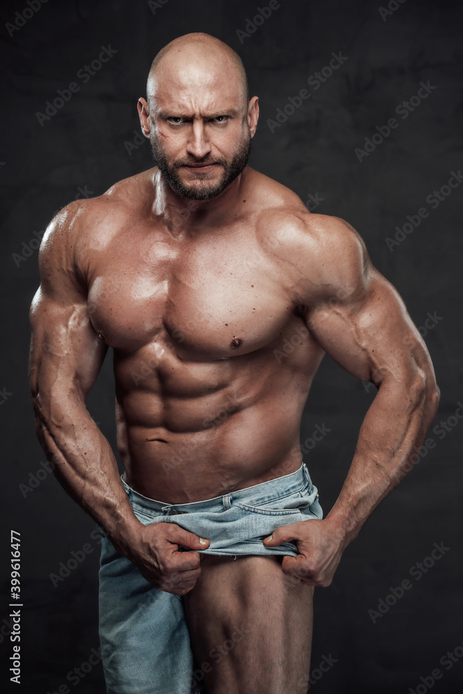 Caucasian muscular man with hairless head and naked torso pulls his shorts showing his muscular leg looking at camera with serious face in dark background.