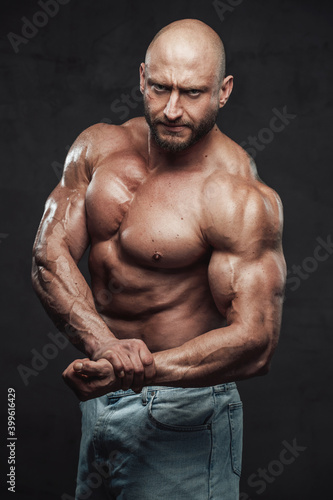 Bearded and hairless man with powerful and muscular build in jeans shorts poses with angry face in dark background looking at camera.