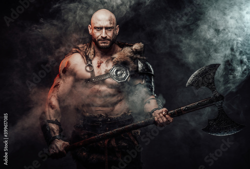 Atmospheric portrait of a powerful and hairless scandinavian warrior posing in dark and smokey background holding his steel two handed axe looking at camera.