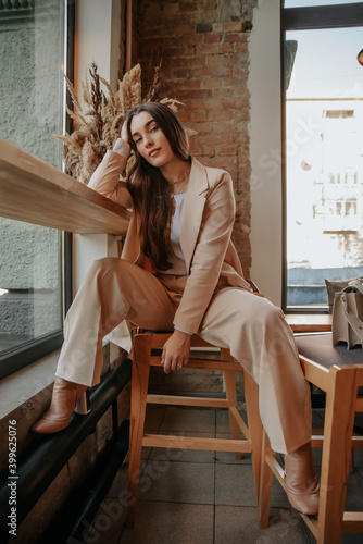 a girl stylist in a beige suit sits with coffee at a huge window in the loft style. long dark hair, charming cocky girl