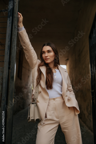 a girl stylist in a beige suit stands in the courtyard with an arch, brooding and bright. long dark hair, charming cocky girl