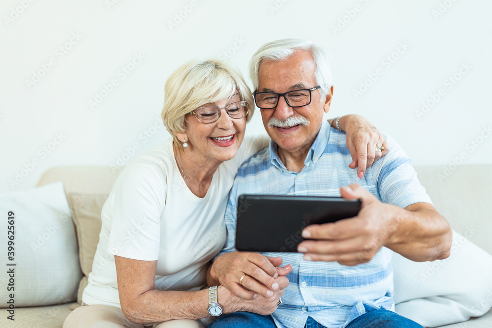 Couple of seniors smiling and looking at the same tablet hugged on the sofa - indoor, at home concept - caucasians mature and retired man and woman using technology - lockdown and quarantine lifestyle Couple of seniors smiling and looking at the same tablet hugged on the sofa - indoor, at home concept - caucasians mature and retired man and woman using technology - lockdown and quarantine lifestyle