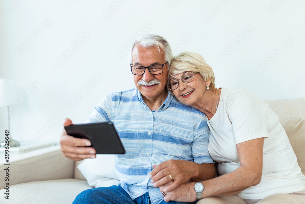 Happy older couple using computer tablet together at home, excited mature man and woman looking at mobile device screen, shopping or chatting online, sitting on cozy couch in living room Happy older couple using computer tablet together at home, excited mature man and woman looking at mobile device screen, shopping or chatting online, sitting on cozy couch in living room