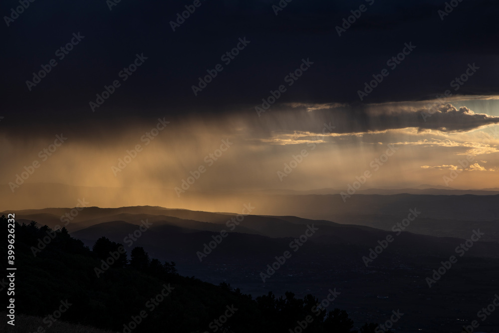 From the top of a mountain, there is a wide view during sunset time (golden hour). The gray clouds full of rain collide with the sunlight creating suggestive contrasts with plays of light.