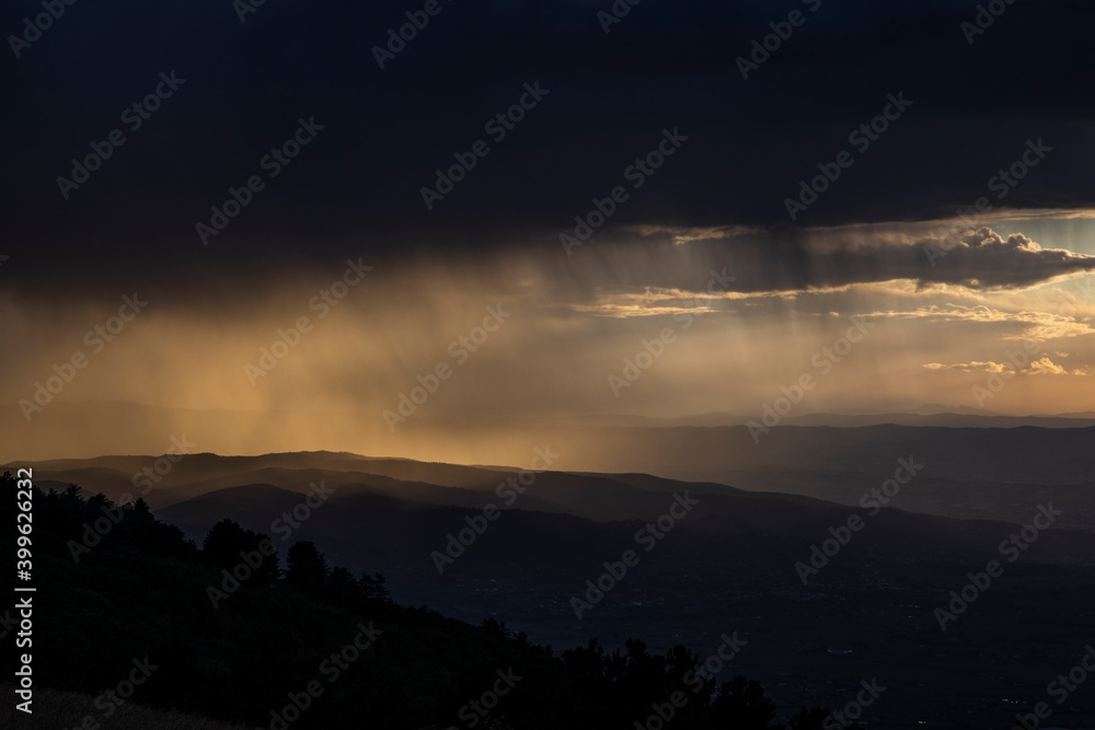From the top of a mountain, there is a wide view during sunset time (golden hour). The gray clouds full of rain collide with the sunlight creating suggestive contrasts with plays of light. From the top of a mountain, there is a wide view during sunset time (golden hour). The gray clouds full of rain collide with the sunlight creating suggestive contrasts with plays of light.