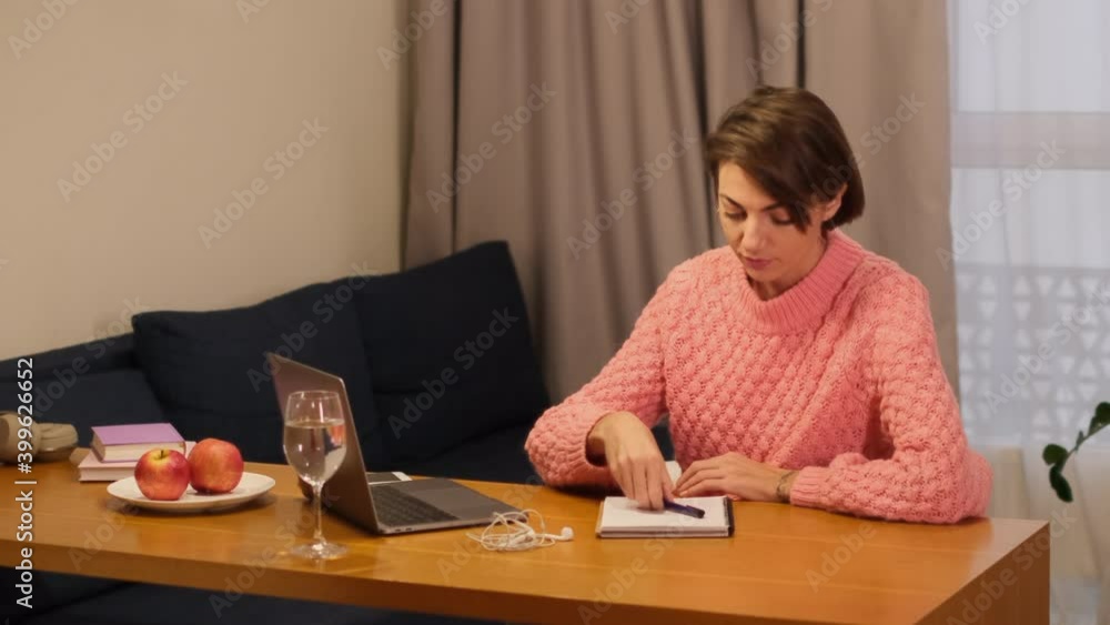 Woman studies at home works writes something in a notebook, looks at a laptop Woman studies at home works writes something in a notebook, looks at a laptop