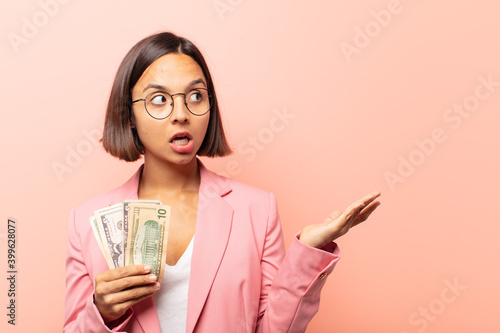 young hispanic woman looking surprised and shocked, with jaw dropped holding an object with an open hand on the side photo