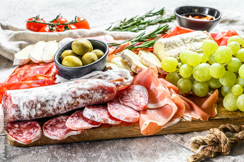 Italian antipasto, wooden cutting board with prosciutto, ham, parma, goat and Camembert cheese, olives, grapes. antipasti. Gray background. Top view