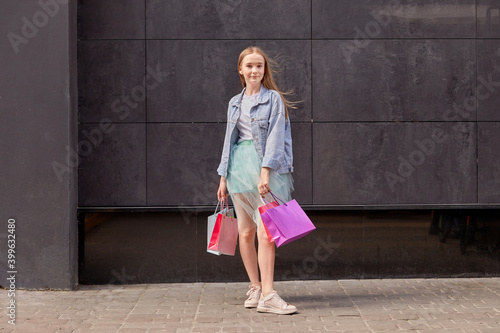 A young girl in jeans and a skirt with shopping in her hands in full growth. A large gray concrete wall. © Evgenia