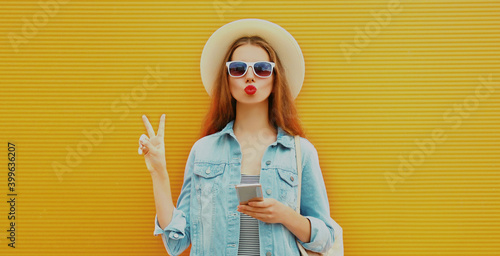 Portrait of young woman with phone blowing lips wearing a summer straw hat over an orange background