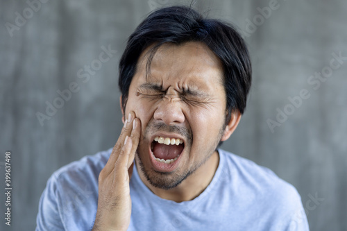 young asian man hands pressed to cheek as he is experiencing severe toothache on gray background