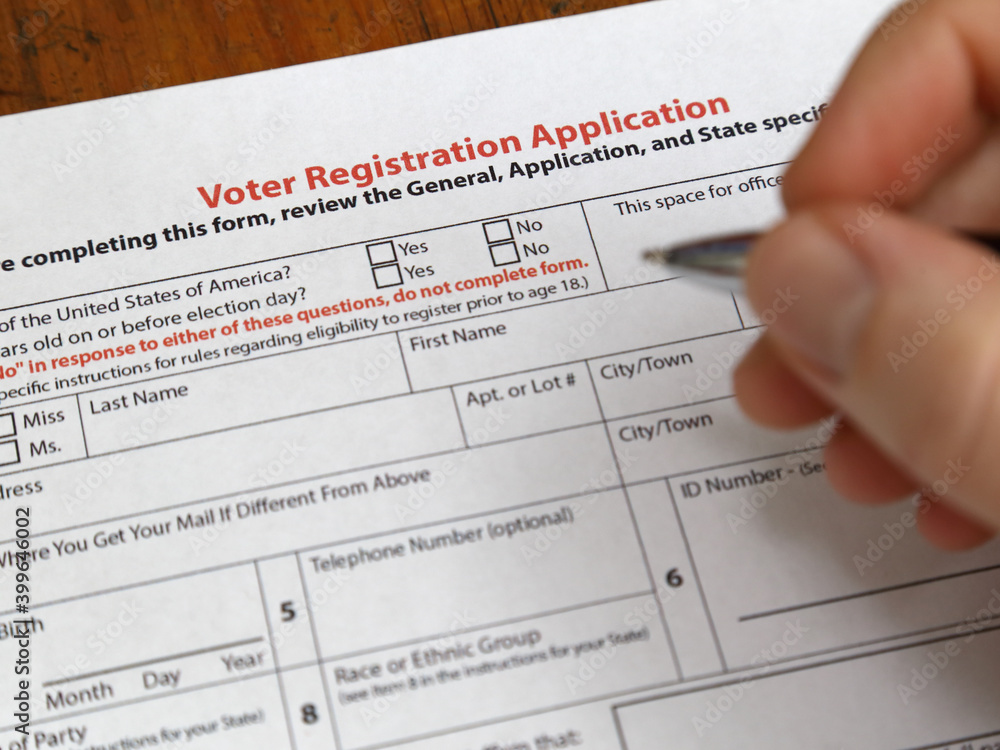 A voter registration form paper copy in the USA is shown up close, with a hand holding a pen about to fill out the sheet. A voter registration form paper copy in the USA is shown up close, with a hand holding a pen about to fill out the sheet.