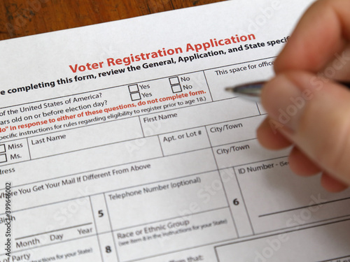 A voter registration form paper copy in the USA is shown up close, with a hand holding a pen about to fill out the sheet.