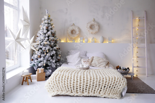 New Years bright interior bedroom with decorated Christmas tree with garlands, white bed, knitted blanket, stairs, stars on window. Christmas interior. Scandinavian style. Location for photo shoot.