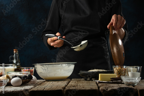 Pastry chef in black uniform pours dough into waffle maker machine for preparing sweet waffle on rustic wooden table with ingredients on dark blue. Frozen motion. Dessert from recipe book.