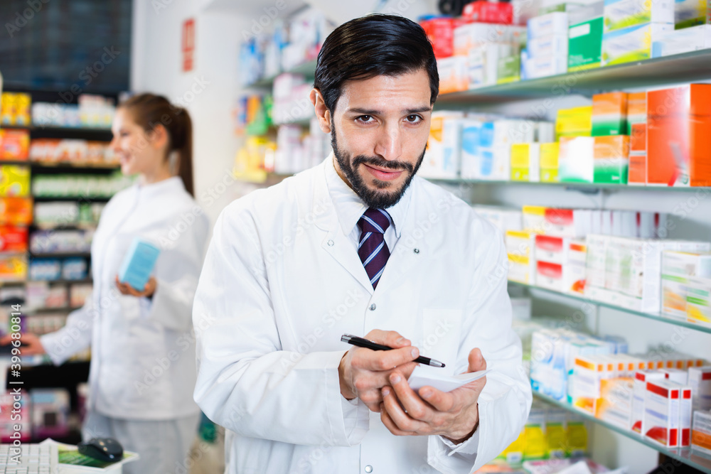 Diligent cheerful smiling male specialist is attentively stocktaking medicines with notebook near shelves in pharmacy.