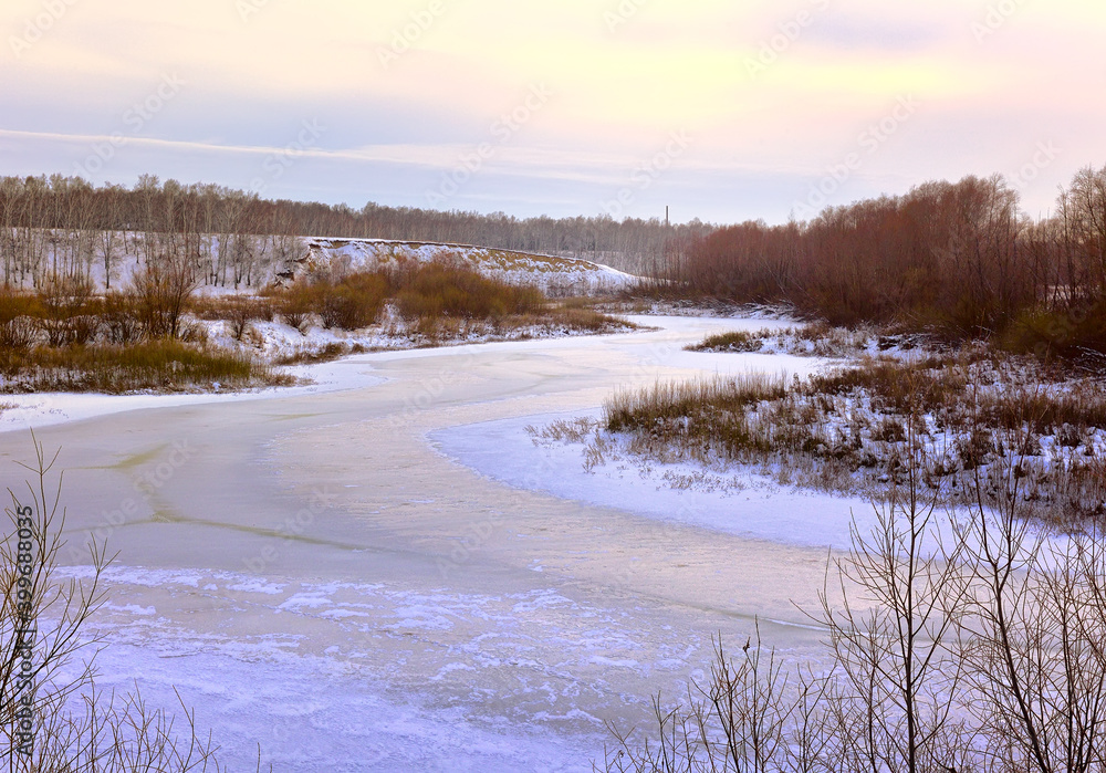 Winter evening over the Tula river. A winding riverbed covered with ice, bare trees in the distance on a snowy Bank Winter evening over the Tula river. A winding riverbed covered with ice, bare trees in the distance on a snowy Bank