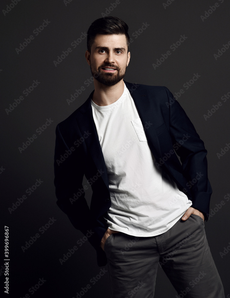 Smiling bearded well-groomed man in jacket, t-shirt and jeans stands with hands in pockets swaying from side to side looking at camera over dark background. Stylish business casual style for men Smiling bearded well-groomed man in jacket, t-shirt and jeans stands with hands in pockets swaying from side to side looking at camera over dark background. Stylish business casual style for men