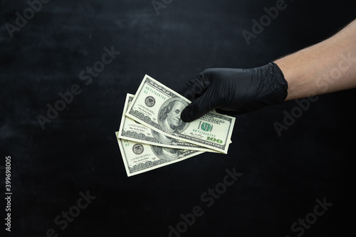 A man's hand in black medical gloves holds three hundred dollar bills in his hand