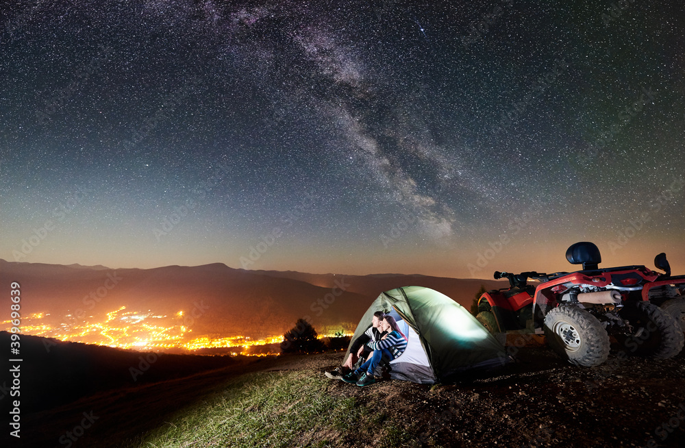 Happy couple man and woman tourists sitting in tourist tent on the top of mountain with atv quad motorbike, enjoying beautiful view of night sky full of stars, Milky way, luminous city on background - obrazy, fototapety, plakaty 