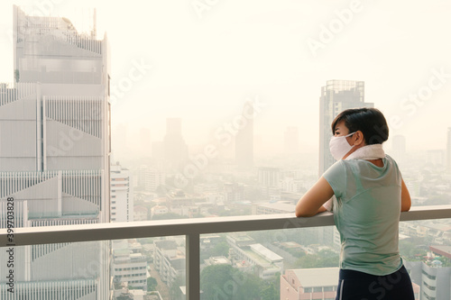 Beautiful Asian woman wear medical face mask look at downtown skyscrapers of city covering with smog, dusk, high PM 2.5 air pollution in serious and dangerous level to health. Air quality problems