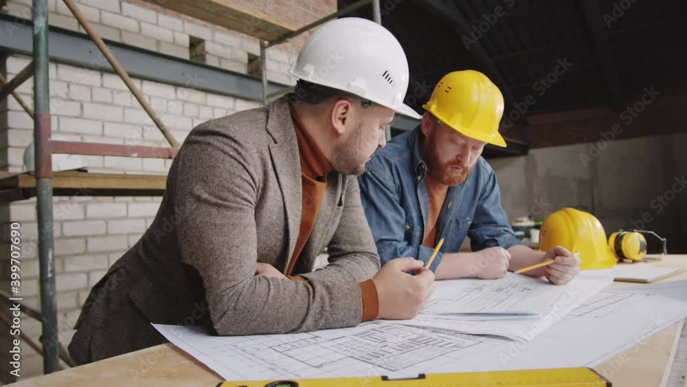 Two Caucasian male engineers in hardhats leaning on worktable and talking about floor plan on paper while working together on project at construction site