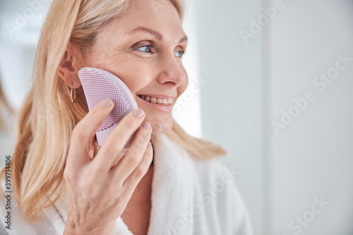 Obraz na plátne Stylish blonde Caucasian female with eclectic facial massager in hand in bathroo