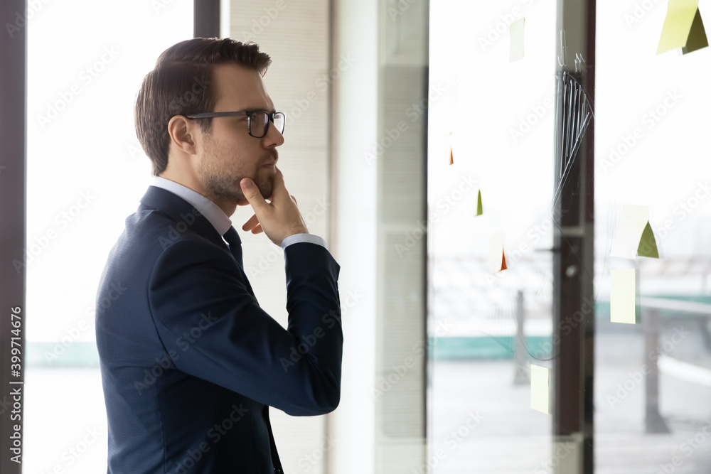 Thoughtful young Caucasian businessman look at glass wall work brainstorm using sticky pads developing business ideas. Pensive male worker do creative thinking in office. Startup, strategy concept. Thoughtful young Caucasian businessman look at glass wall work brainstorm using sticky pads developing business ideas. Pensive male worker do creative thinking in office. Startup, strategy concept.