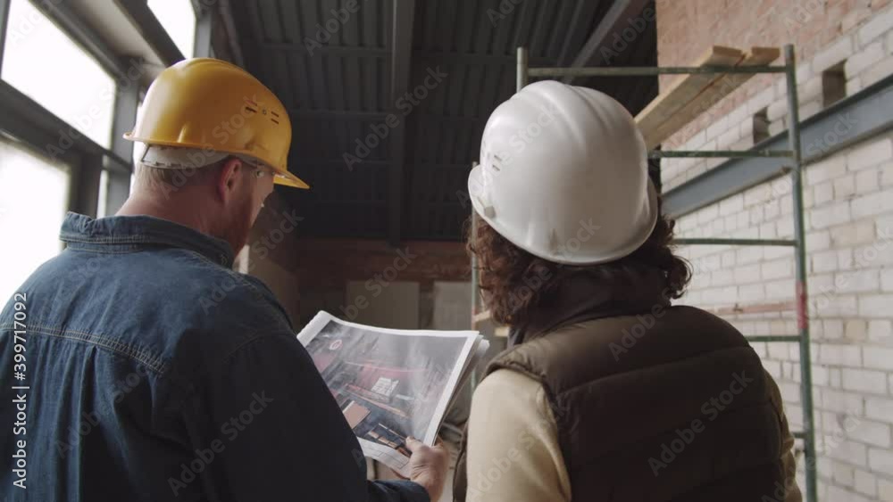 Back view of male and female engineers in hardhats standing in unfinished apartment and discussing paper slides of construction project Back view of male and female engineers in hardhats standing in unfinished apartment and discussing paper slides of construction project