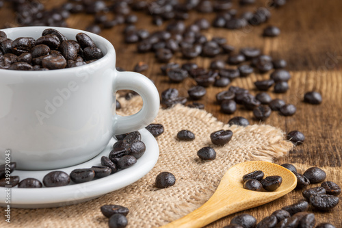 Close-up of white cup and plate with coffee beans, on burlap, with wooden spoon and more beans on wooden table, selective focus, horizontal