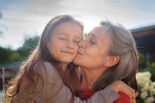 Senior grandmother with gray hair wearing red sweater with her little granddaughter are hugging in the garden and during sunny day outdoors, mother's day