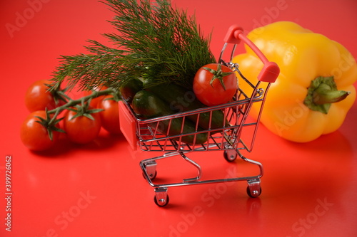 Close up of supermarket grocery push cart for shopping with cucumbers , tomatoes, bell pepper  and dill on red background. Concept of shopping. Copy space for advertisement. photo