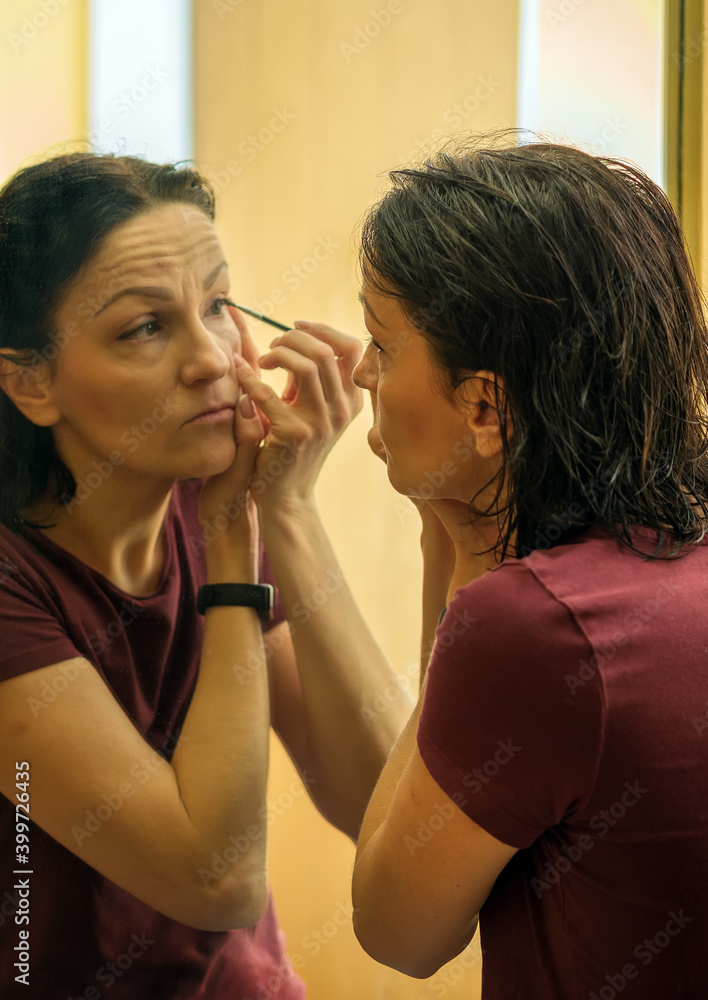 Naklejka premium Middle age Caucasian woman applying makeup in front of a large mirror.